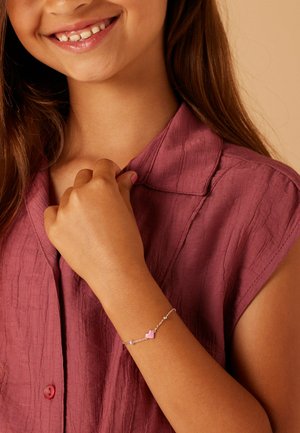 Silbernes Armband mit einem rosa Herz-Anhänger, getragen an einem braunen Arm, mit einem strukturierten burgunderfarbenen Hemd im Hintergrund.