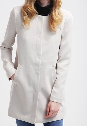 Frau mit welligem blondem Haar, die einen strukturierten, cremefarbenen langen Mantel, darunter ein schwarzes Oberteil und blaue Jeans trägt, mit den Händen in den Manteltaschen.