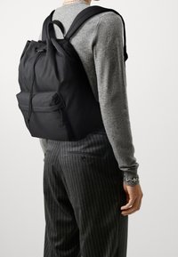 Sac à dos noir avec cordon de tirage, présentant un extérieur lisse, une poche zippée à l'avant et des bretelles ajustables, porté sur un tissu gris.