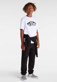Vit grafisk t-shirt med "VANS OFF THE WALL" logotyp, svarta sweatpants och svart-vita rutig slip-on skor. Avslappnad outfit.