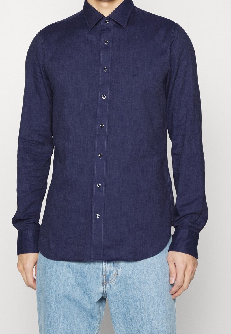 Homme portant une chemise bleu foncé à manches longues, boutonnée avec col, et un jean bleu clair, debout devant un fond clair uni.