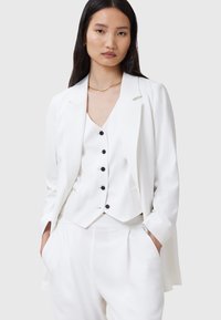 Weißes maßgeschneidertes Blazer-Jackett über einem weißen Unterhemd mit schwarzen Knöpfen, kombiniert mit passenden Hosen. Glattes Material mit strukturierter Passform und minimalistischem Design.