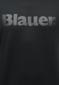 Μαύρο ύφασμα με μεγάλα, παχιά, γκρίζα γράμματα "Blauer." τοποθετημένα οριζόντια και κεντραρισμένα σε όλη την επιφάνεια.