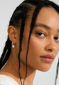 Boucle d'oreille en argent avec un charme floral, présentée sur une oreille. Des cheveux tressés complètent le design de la boucle d'oreille sur un fond neutre.