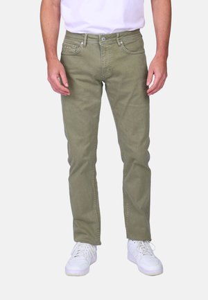 Homme portant un jean slim vert olive et des baskets blanches, debout devant un fond uni avec les mains relâchées le long du corps.