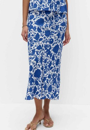 Kobieta ubrana w niebiesko-białą, kwiatową, midi spódnicę oraz beżowe sandały na obcasie, stojąca na jednolitym białym tle.