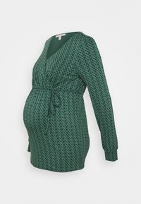 Zelený těhotenský top s oblíbeným zavinovacím designem, dlouhým rukávem a zavazovací páskou v pase. Obsahuje texturovaný vzor malých diamantů po celém povrchu.