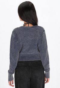 Maglione cropped blu navy con lavorazione a maglia testurizzata, polsini e scollatura a costine. Indossato con pantaloni neri a vita alta, che mostrano il design posteriore.