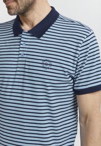 Tamsiai mėlynas ir šviesiai mėlynas dryžuotas polo marškinėliai. Yra klasikinis apykaklė, trys mygtukai ir siuvinėtasis logotipas ant krūtinės. Minkšta medžiaga.