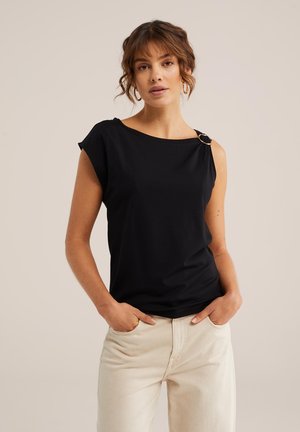 Femme aux cheveux ondulés portant un haut noir sans manches avec un détail en anneau sur l'épaule et un pantalon beige clair, les mains dans les poches.