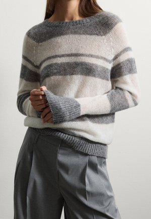 Personne portant un pull en laine doux rayé blanc et gris, associé à un pantalon plissé gris ample, debout contre un mur uni.