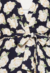 Květinové šaty přes hlavu mají námořnické pozadí s krémovými růžemi a zelenými listy, představující výstřih do V a detail s páskem na zavazování.