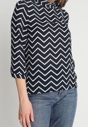 Vrouw draagt een zwart-wit zigzagpatroon lange mouwen top met 3/4 mouwen en een blauwe spijkerbroek, staand tegen een effen achtergrond.