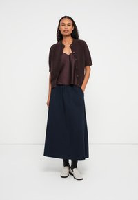 Cardigan curto bordô de mangas curtas sobre uma blusa roxa escura, combinado com uma saia longa plissada azul-marinho e sapatos de slip-on em tom claro.