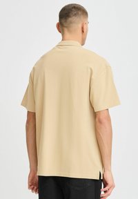 Beige kortermet skjorte med avslappet passform, krage og sidesplitter. Materialet virker lett og glatt med et enkelt design.