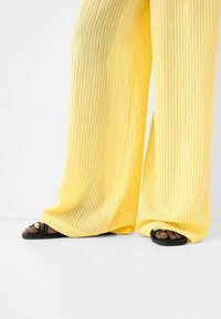 Gula plisserade byxor med vida ben och vita ränder. Svarta sandaler med ett cirkulärt accent som visar fram tårna. Minimalistisk och lätt design.