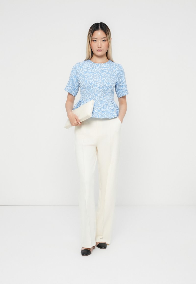 Hellblaues, floral gemustertes kurzärmliges Oberteil, weiße, maßgeschneiderte Hose, hält eine cremefarbene Clutch, mit schwarz-beigen zweifarbigen Schuhen.