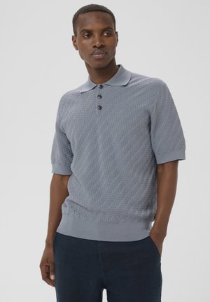 Mand iført lyseblå struktureret kortærmet poloshirt med tre mørke knapper og mørkeblå bukser, stående med den ene hånd i lommen.