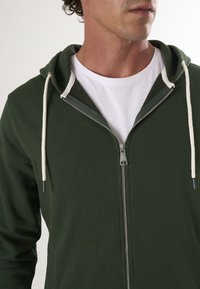 Groene zip-up hoodie met een witte trekkoord, voorzien van een ritssluiting en een textuur binnenvoering, gecombineerd met een wit t-shirt eronder.