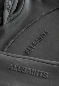 Harmaa huppari, jossa on kohotettu "ALLSAINTS"-logo. Ominaisuuksiin kuuluu pehmeä pinta, näkyvät saumat ja huppu, jossa on kiristysnyöri.