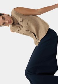 Tan mouwloos topje met twee borstzakken en donkere knopen, gecombineerd met marineblauwe wijdpantalon met een gladde textuur en strakke lijnen.