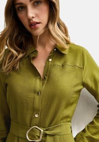 Olivgrön skjorta med knappar och krage, med subtil textur och ett midjebälte med spänne i silvermetal.