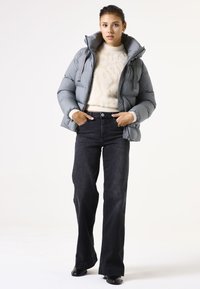 Grijze gewatteerde jas met een hoge kraag, gecombineerd met een crèmekleurige trui en donkere flared jeans. Model draagt zwarte schoenen. Glad textuur van de stof.