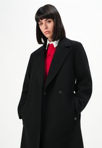 Donna con capelli scuri e lisci che indossa un soprabito nero sopra un maglione rosso e una camicia bianca con colletto, in piedi con le mani in tasca.