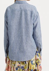 Kofta med långa ärmar i bomull med blå och vita vertikala ränder, ryggveck, knappmanchetter och klassisk krage, som bärs över en blommig kjol.