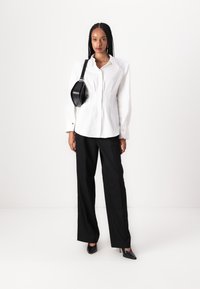 Blusa blanca con hombros estructurados y botones dorados, combinada con pantalones anchos negros y zapatos de tacón de punta negra. Bolso cruzado negro.
