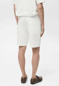 Shorts en coton blancs avec une coupe droite, doté d'une poche arrière, associés à des chaussures brunes, vus de l'arrière.