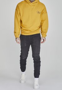 Gelbes Kapuzensweatshirt mit einem Logo auf der Vorderseite, kombiniert mit schwarzen Slim-Fit-Jogginghosen mit Seitentaschen und gerippten Säumen. Weiße Sneaker.