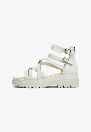 Sandales blanches à lanières avec plusieurs lanières étroites, une semelle épaisse et un talon arrière, présentant un détail de boucle sur les lanières de la cheville.