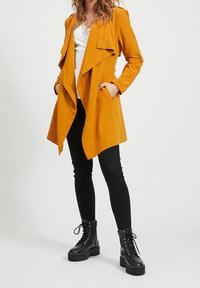 Musztardowożółty trencz z otwartym przodem, oversize'owymi klapami i bocznymi kieszeniami, w zestawie z czarnymi obcisłymi dżinsami i czarnymi botkami na sznurówki.