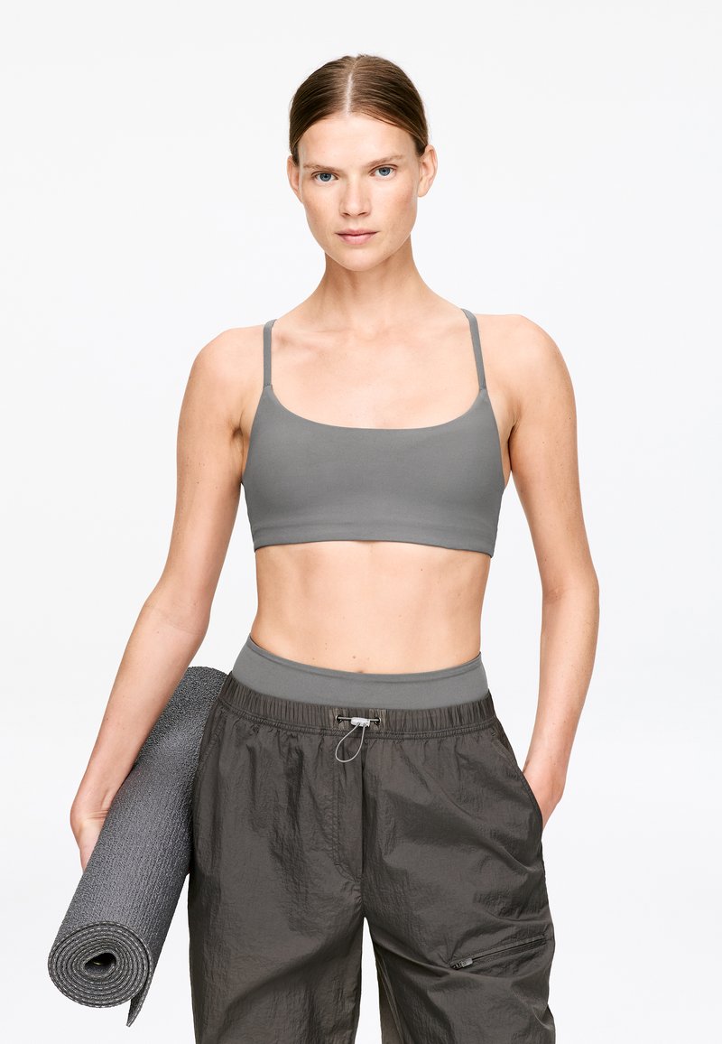 Brassière de sport grise à fines bretelles, associée à un pantalon gris foncé léger avec taille élastique. Le sujet tient un tapis de yoga roulé.