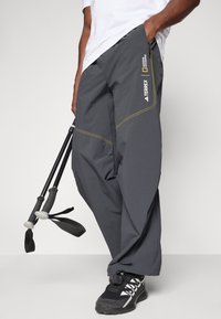Pantaloni impermeabili grigi con dettagli di cucitura gialla, vita regolabile e tasche laterali, abbinati a scarpe da trekking nere e bastoni da trekking.