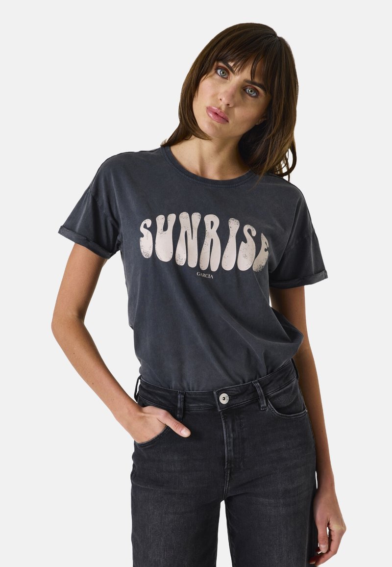 Graues Baumwoll-T-Shirt mit aufgerollten Ärmeln, das mit einem auffälligen weißen "SUNRISE"-Grafik versehen ist. Kombiniert mit hochtaillierten schwarzen Jeans.