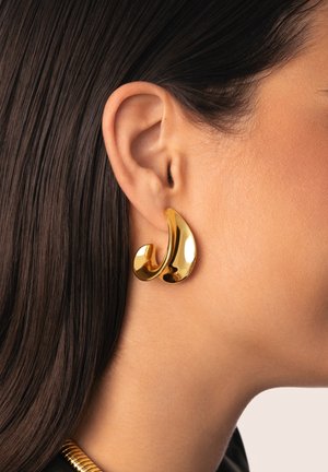 Boucles d'oreilles en ton or avec une forme fluide en goutte d'eau. La surface brillante reflète la lumière, mettant en valeur une texture lisse et un design élégant.