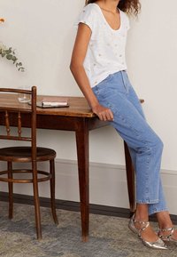 Vit kortärmad topp med metalliska detaljer, ljusblå jeans och silverfärgade ballerinaskor, placerad vid ett träbord med ett glas och en anteckningsbok.