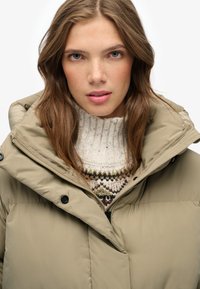 Chaqueta de plumas color caqui con un collar alto, que presenta una cremallera frontal y botones a presión, superpuesta sobre un suéter de punto crema con acentos multicolores.