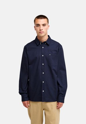 Jeune homme portant une chemise bleu marine à boutons avec une poche avant et un pantalon beige, debout devant un fond clair uni.