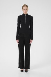 Zwarte ribbelgebreide top met hoge kraag en zilveren rits, gecombineerd met loszittende zwarte broek en puntige zwarte schoenen.