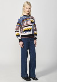 Pull en tricot avec des motifs géométriques colorés en bleu, marron, jaune et noir. Associé à un jean en denim évasé et des bottes noires.