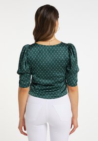 Grøn satin bluse med puffærmer og et geometrisk mønster. Har en rund halsudskæring og en tætsiddende silhuet, parret med hvide bukser.