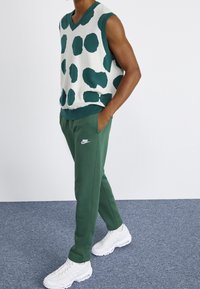 Grön och vit prickig stickad väst med V-ringning, parat med gröna sweatpants och vita sneakers. Avslappnad, atletisk passform och material.