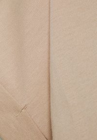 Gros plan sur un tissu en tricot beige montrant une texture détaillée et une petite couture brodée.