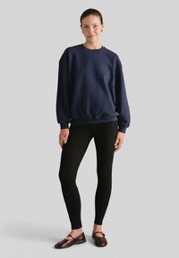 Ciemnogranatowa, oversize bluza z opuszczanymi ramionami, w połączeniu z czarnymi legginsami i ciemnobrązowymi, płaskimi butami. Na jednolitym tle.