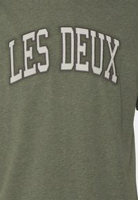 Oliwkowa bawełniana koszulka z nadrukiem "LES DEUX" w białym i ciemnoszarym kolorze, z okrągłym dekoltem i krótkimi rękawami.