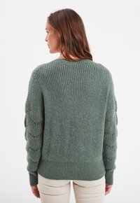 Pull en tricot vert avec texture côtelée, doté de détails ajourés sur les manches et d'une coupe décontractée. L'ourlet est également côtelé pour un détail supplémentaire.