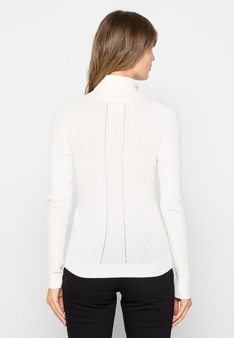 Suéter blanco de manga larga con canalé y cuello alto. Presenta un ajuste ceñido y costuras abiertas en la parte posterior. Textura suave y elementos de diseño mínimos.